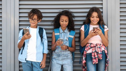 Three children with backpacks standing against a wall, smiling and looking at their smartphones. An image for Haven Online Safety's Student Curriculum a resource for schools.