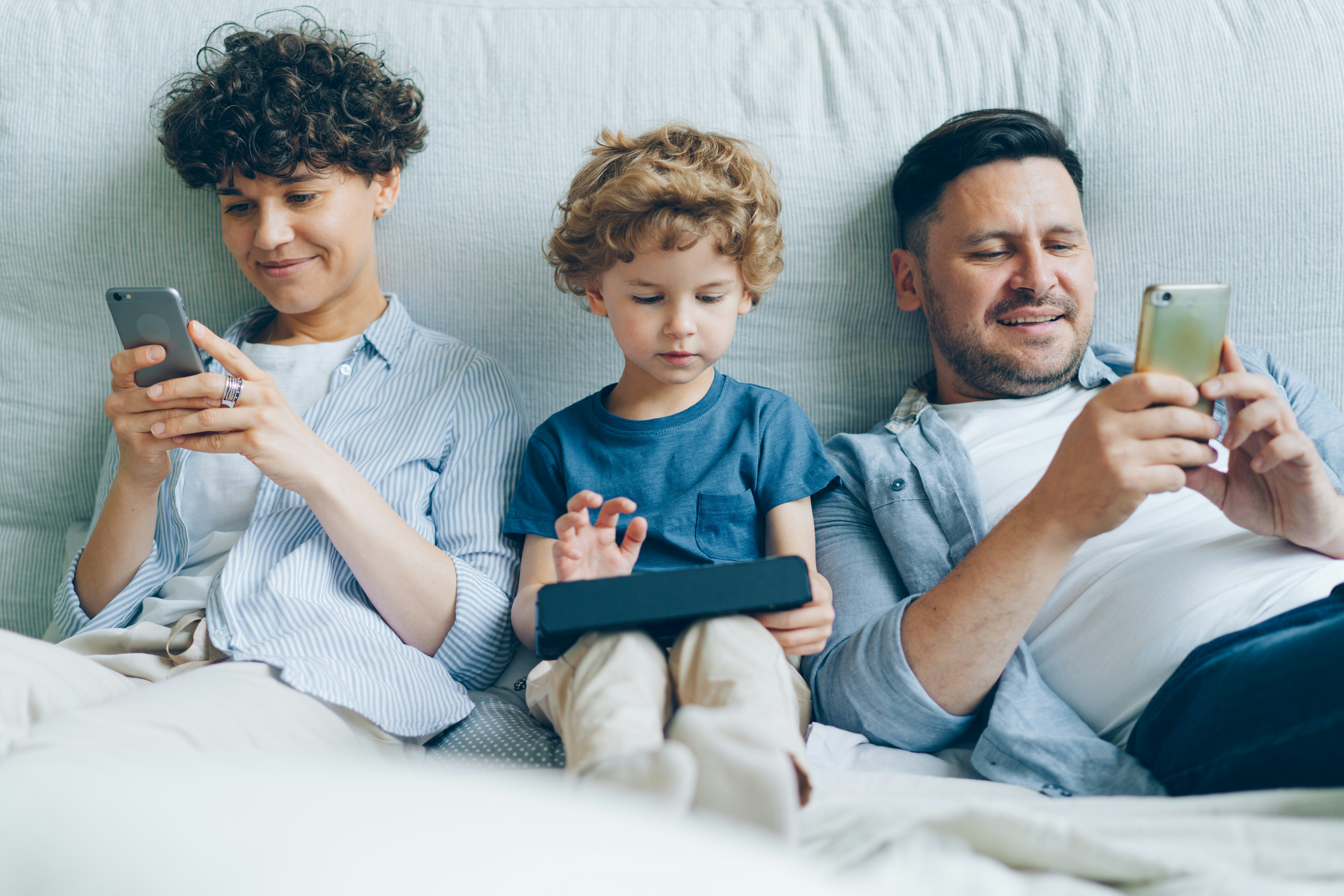 Smiling mother, father, and child lying on bed using smartphones and tablet devices.