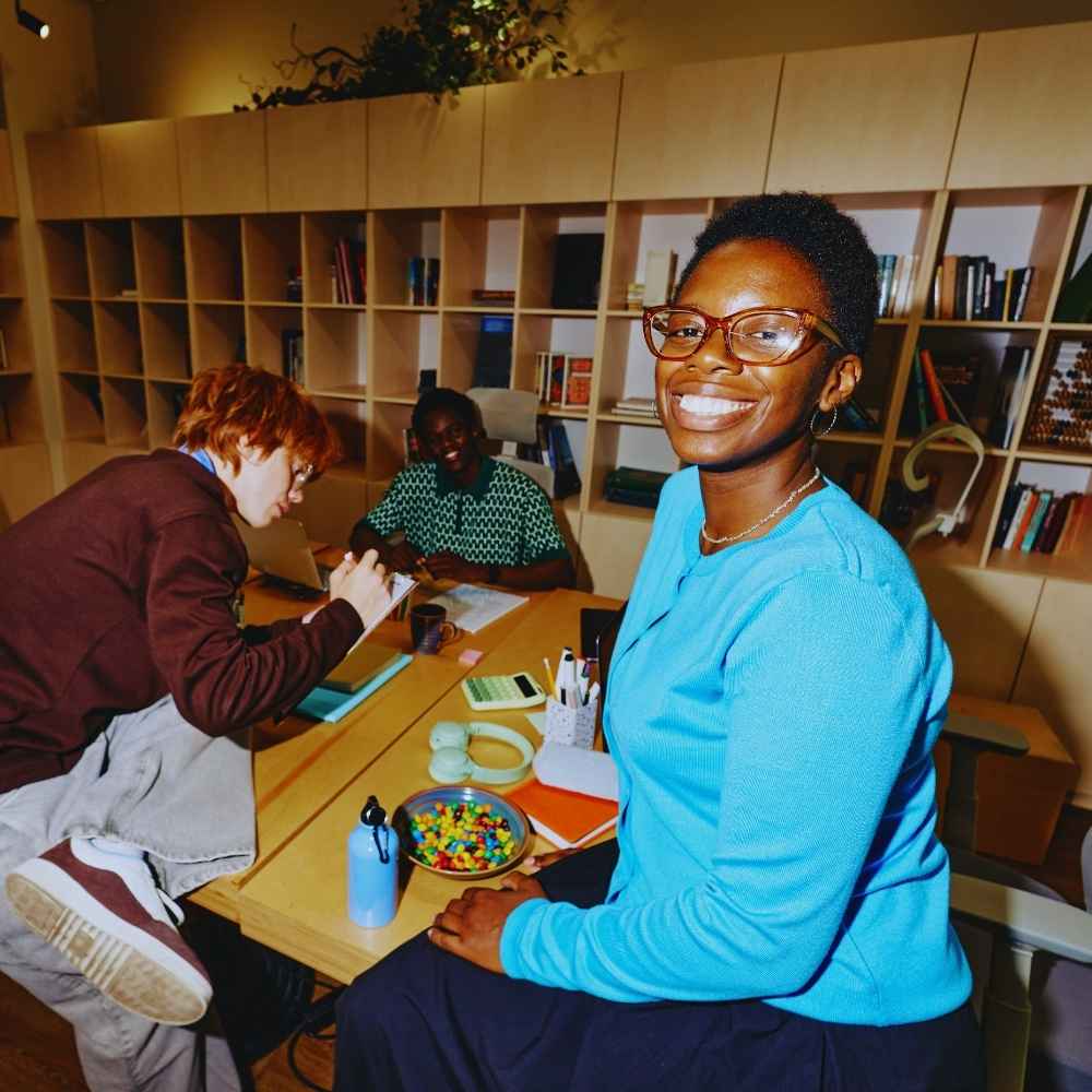 Smiling woman in blue sweater sits at table with two people working in a room with bookshelves.