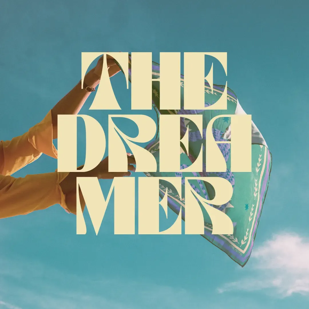 A person holding up a flowing scarf against a blue sky. Large text over the image says: 'THE DREAMER'.