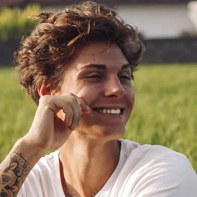 Smiling young man with curly hair and a tattoo on his arm, sitting outdoors in a field, wearing a white t-shirt.