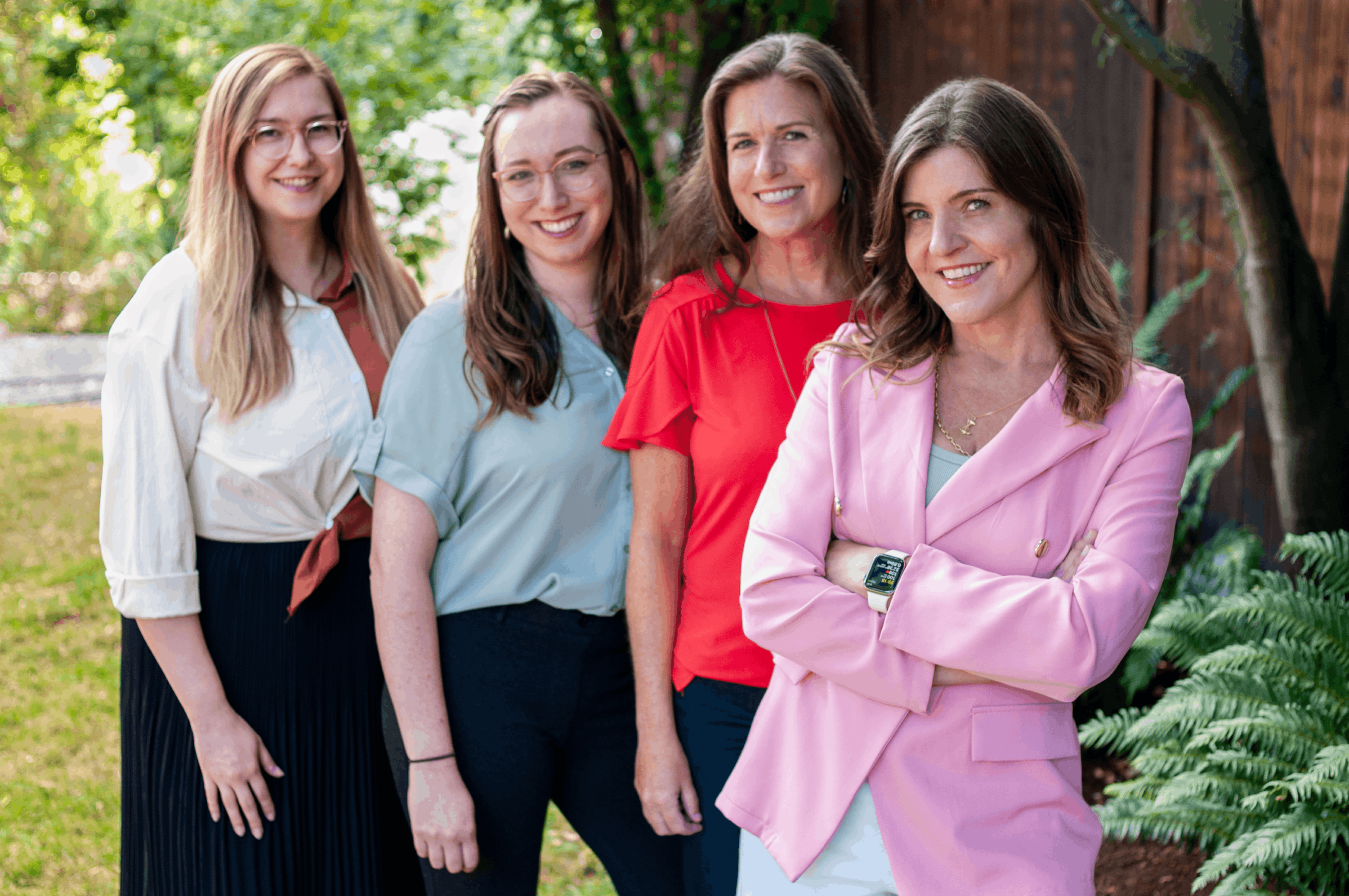 Four smiling women standing outdoors in front of greenery and a wooden fence, dressed in casual and business-casual outfits.