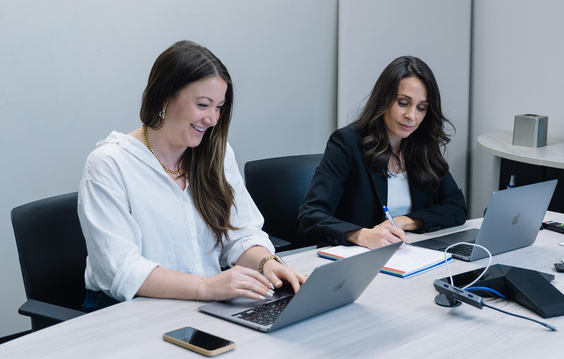 Two women working at a desk with laptops; one is smiling and typing, the other is writing in a notebook.