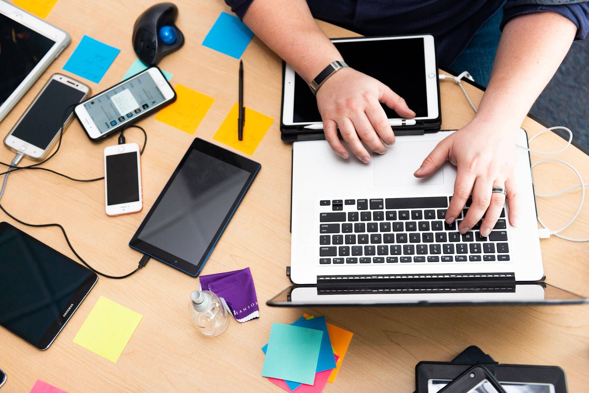 Person typing on a laptop surrounded by various tablets, smartphones, sticky notes, and a mouse on a wooden desk.