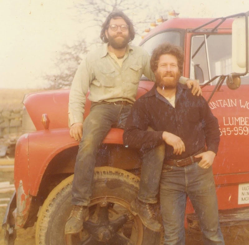 Two men with beards posing beside a red lumber company truck in the 1970s, one sitting on the tire and one standing