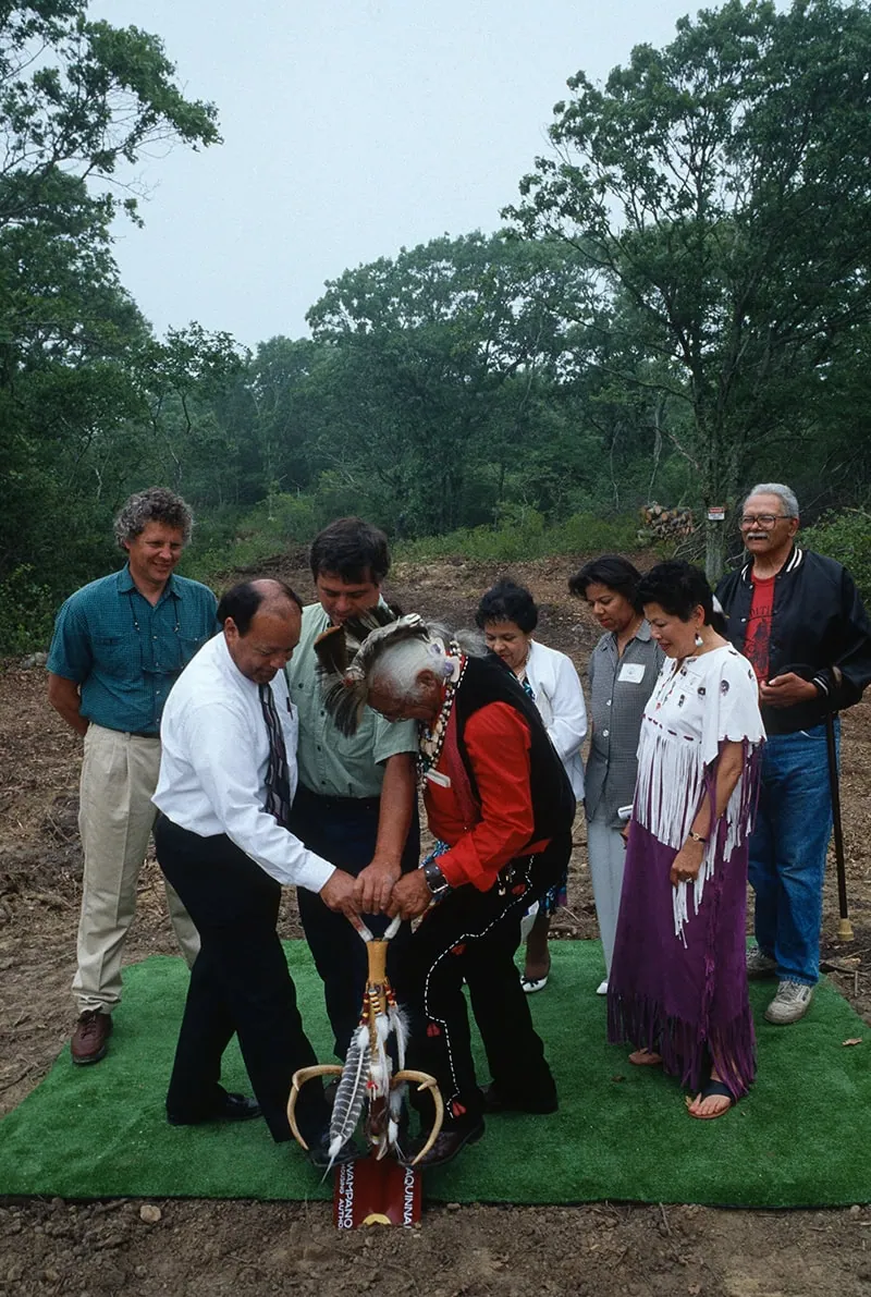 Multicultural group participating in a groundbreaking ceremony, with a Native American elder in traditional red clothing using a ceremonial shovel