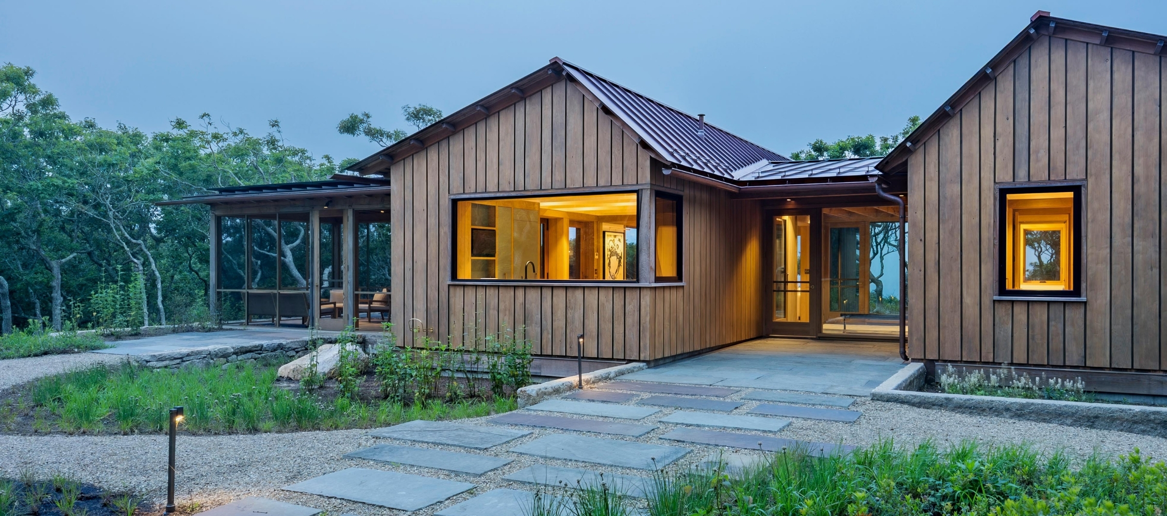 Single story South Mountain house new construction nestled in the landscape overlooking Vineyard Sound, lit from within at dusk.
