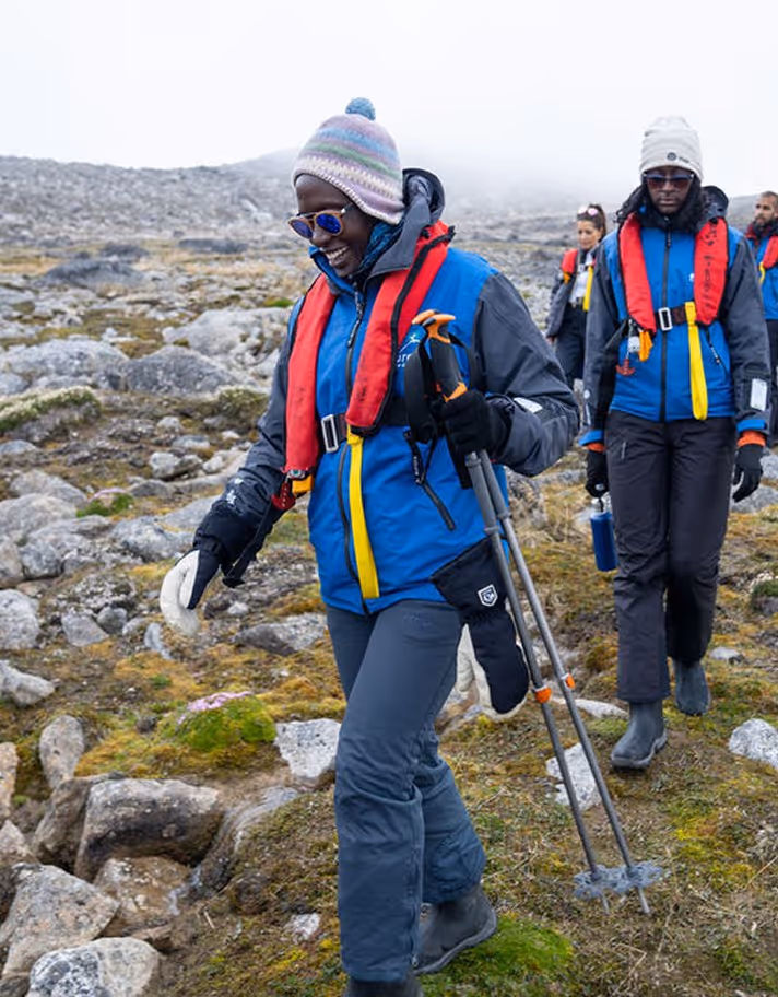 A group of hikers navigating rocky terrain on a cloudy day. One hiker, wearing a blue jacket, grey pants, and a beanie, uses trekking poles while others follow behind, dressed for rugged outdoor conditions.