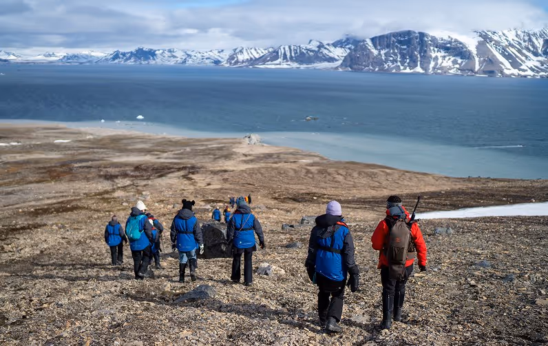 A group of hikers wearing blue jackets walking down a rocky slope towards a glacial body of water, with snow-capped mountains in the background on a clear day.