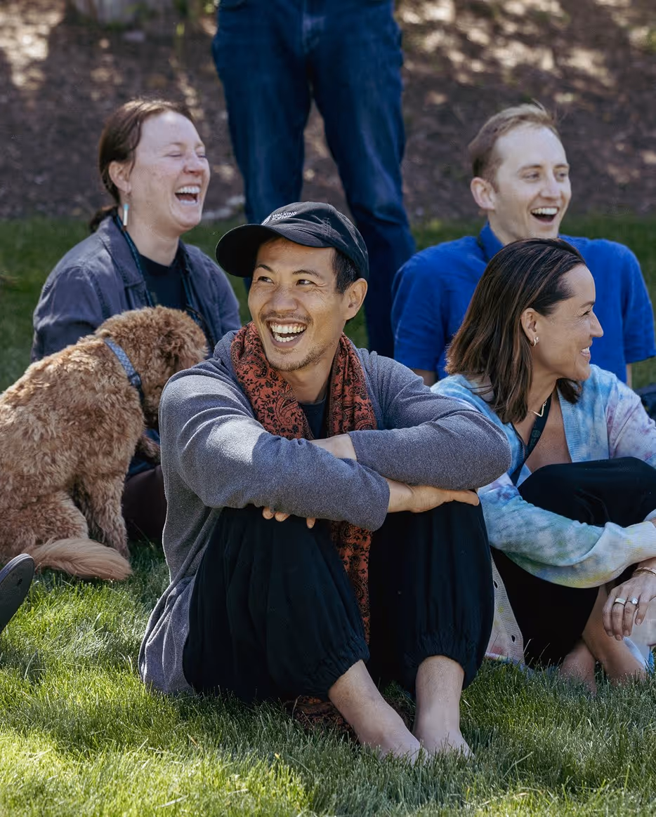 A group of five people sitting on grass, smiling and laughing. One person, a man in a black cap and gray sweater, is sitting cross-legged with a red scarf. A brown dog is partially visible near one of the women.
