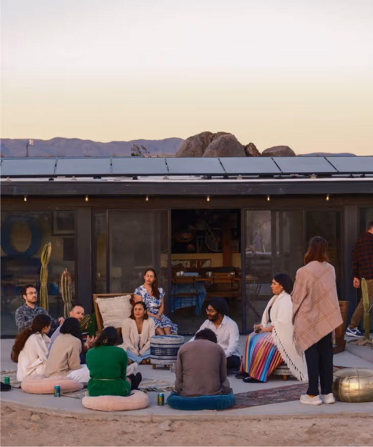A diverse group of people sitting on cushions outdoors near a modern house at sunset, with cacti and mountains in the background.