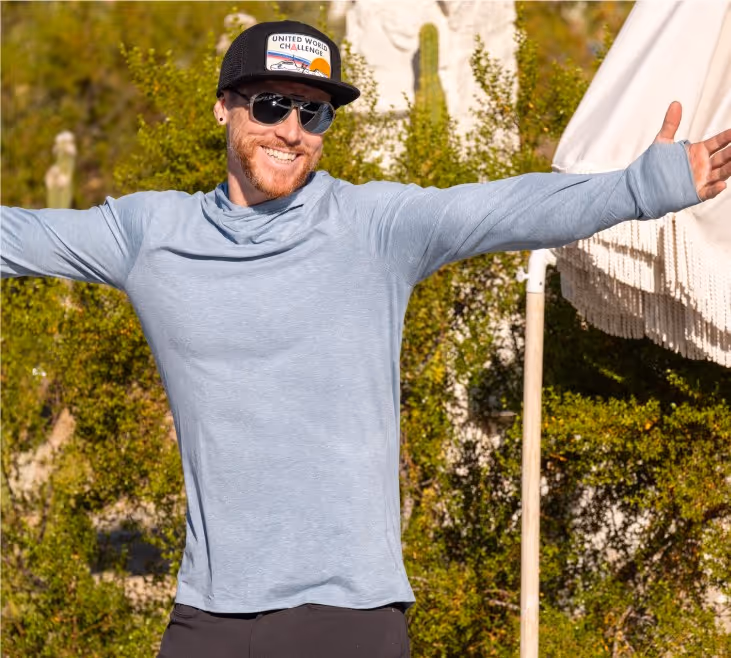 A man wearing a light blue long-sleeve shirt and sunglasses smiles with arms outstretched outdoors, surrounded by greenery and a white umbrella in the background.