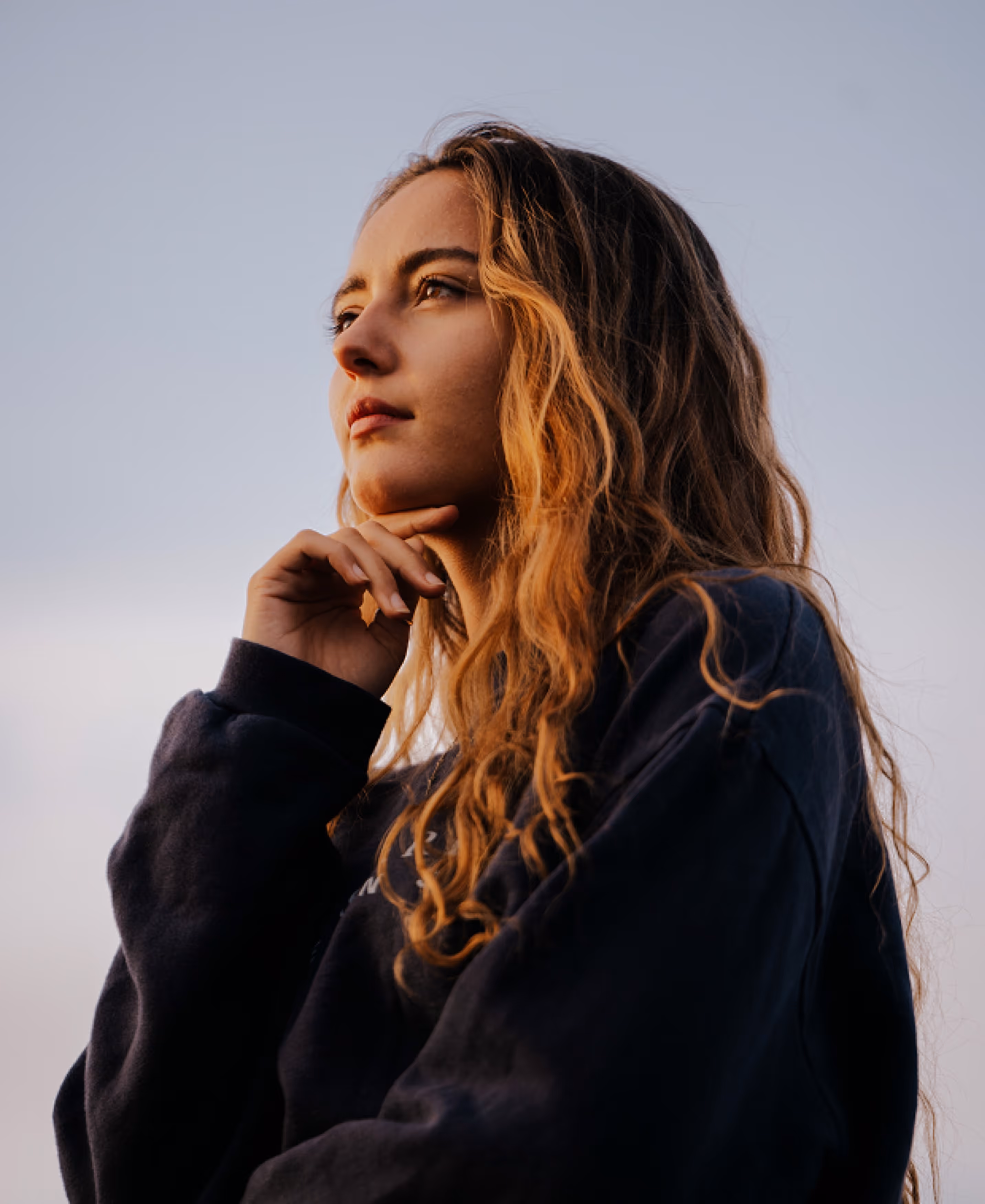 A woman with long, wavy hair stands outdoors, her chin resting on her hand as she gazes thoughtfully into the distance. She is wearing a dark long-sleeve shirt with a soft background.