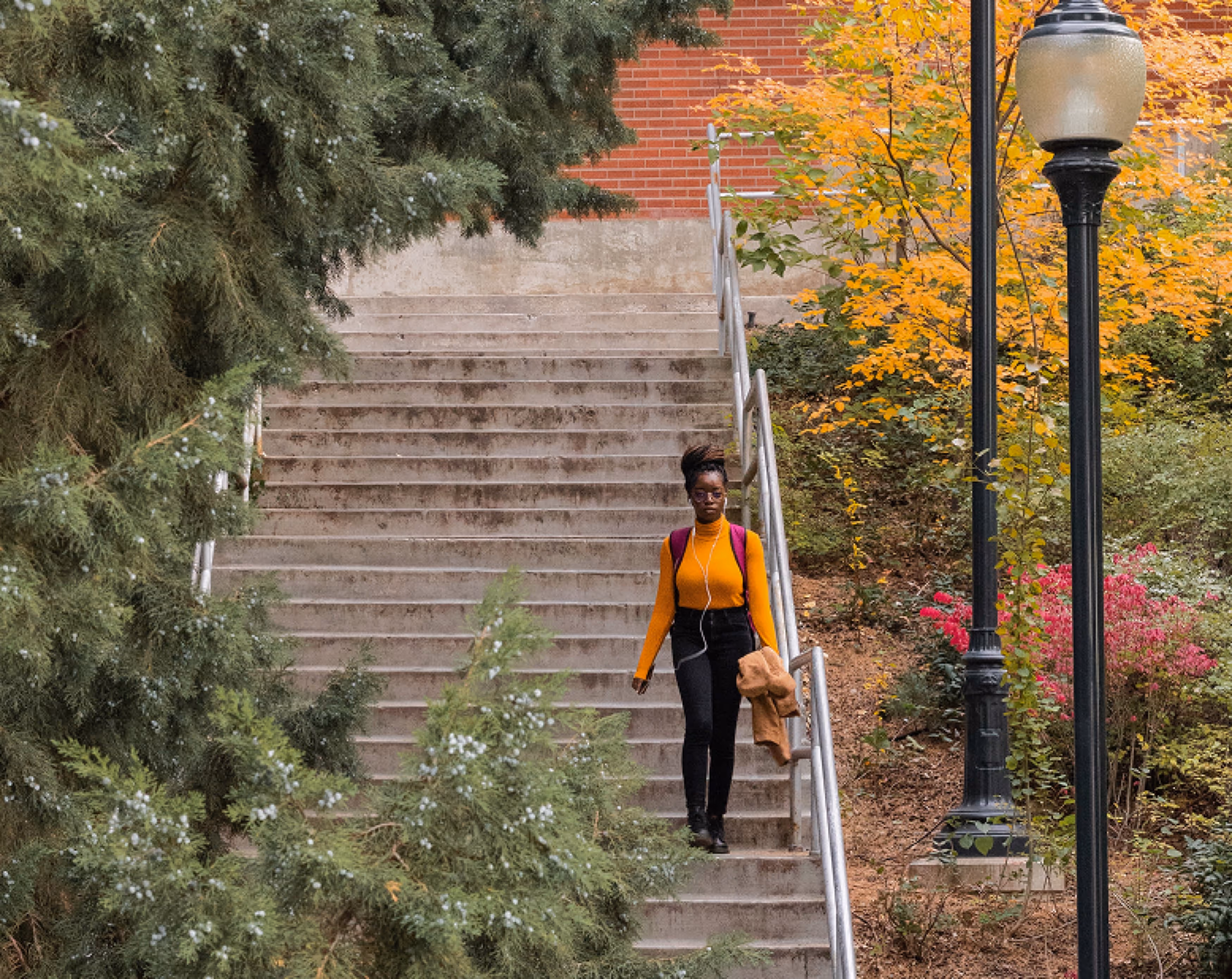 A woman in a bright orange top and black pants walking down outdoor stairs surrounded by trees and colorful autumn foliage. A lamppost is positioned on the right, enhancing the picturesque setting.