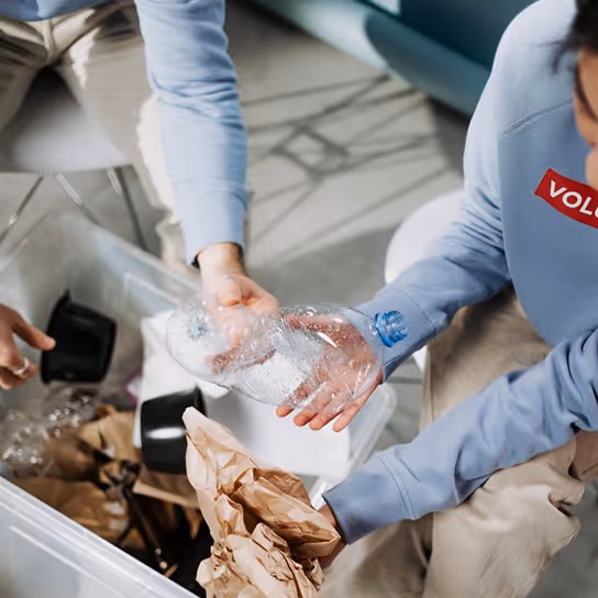 Two individuals sorting recyclable materials in a clear container. One person holds a plastic water bottle, while the other is managing crumpled brown paper. They are engaged in an organized recycling effort.
