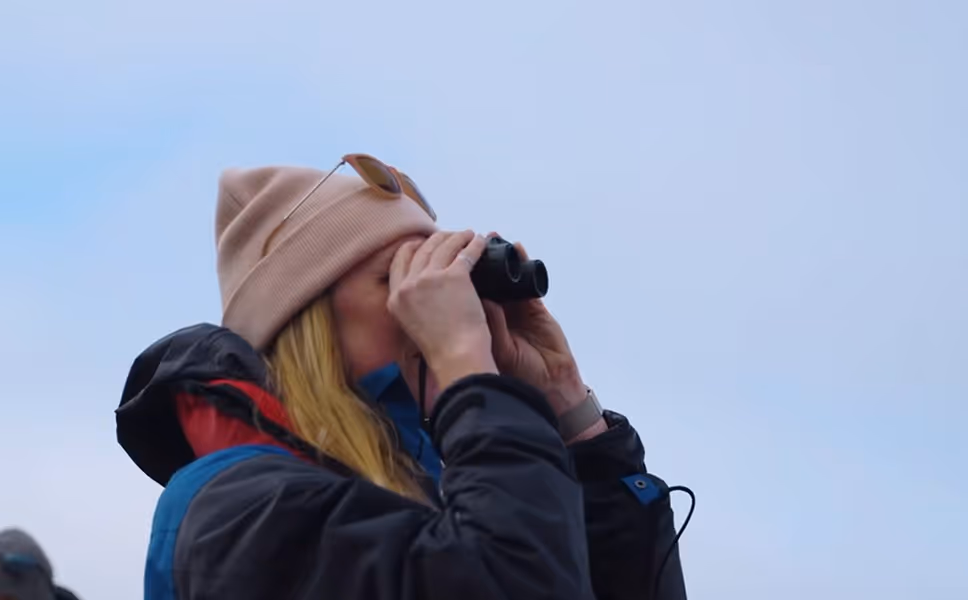 A woman wearing a beige beanie and a jacket stands outdoors, looking through binoculars towards the sky. Her long hair is visible, and there is a soft blue sky in the background.