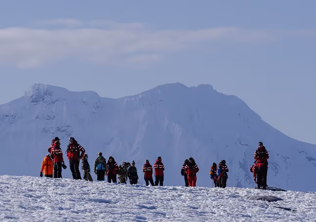 A group of people in colorful winter jackets walking across a snowy landscape with mountains in the background under a clear sky.
