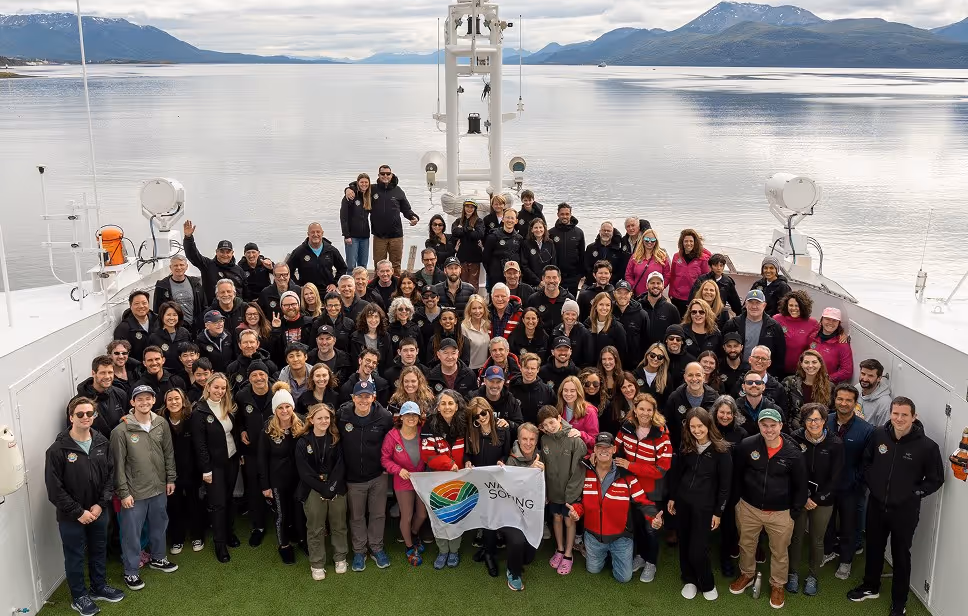 A large group of people posing for a photo on the deck of a ship, with mountains and calm water in the background. Participants wear jackets, some holding a banner with a logo. The scene captures the sense of teamwork and collaboration.