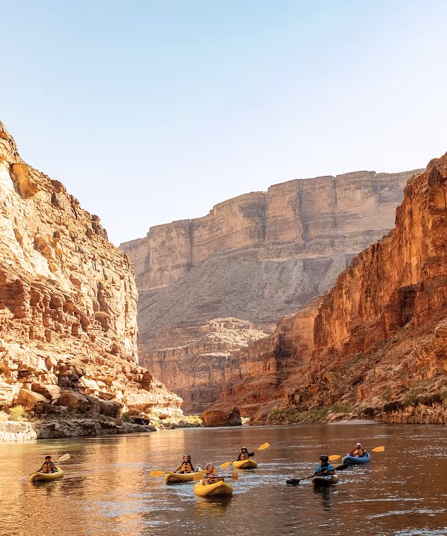 A group of kayakers maneuvering through a river surrounded by large rock formations and cliffs under a clear blue sky.