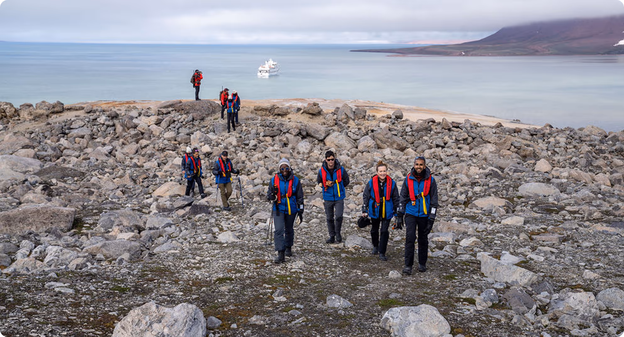 A group of eight hikers wearing vibrant jackets walking on rocky terrain near a shoreline, with a boat visible in the background on calm waters under a cloudy sky.