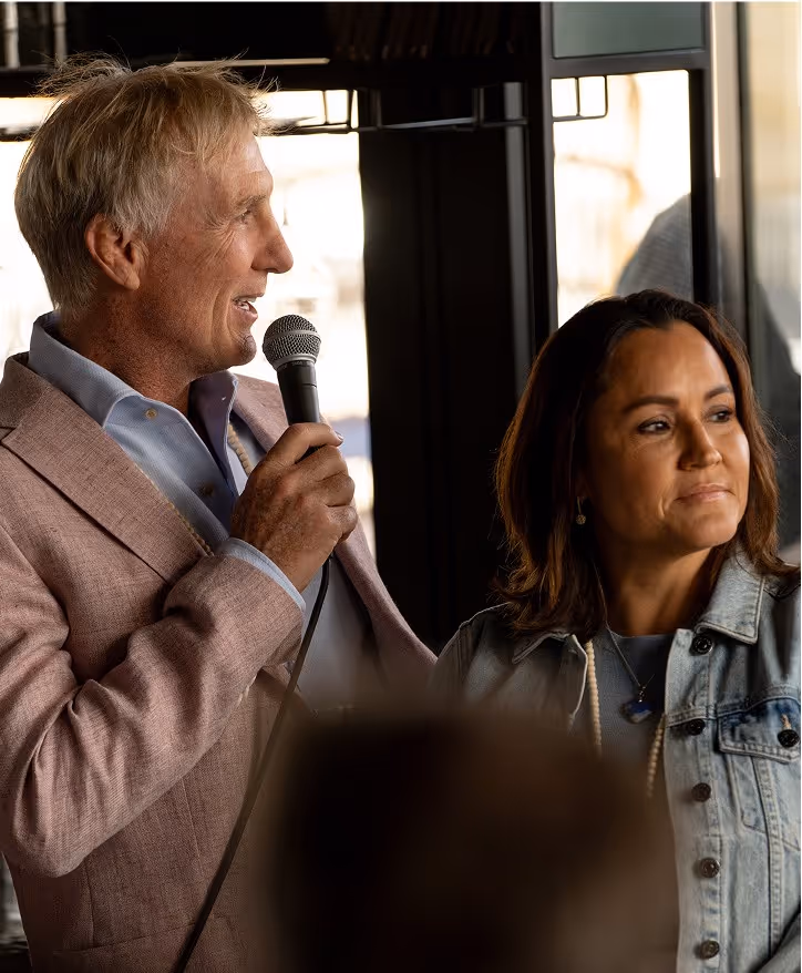 A man in a light pink suit holds a microphone while speaking, with a woman in a denim jacket standing next to him, listening. The scene is set indoors with large windows in the background.
