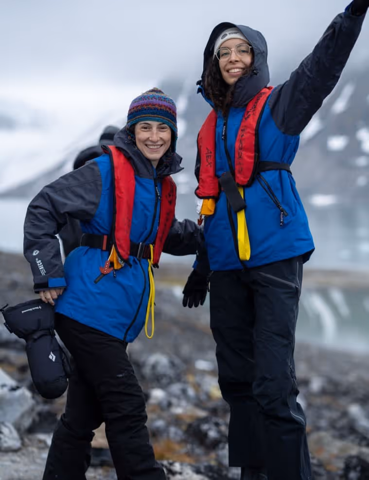 Two women wearing life jackets and outdoor gear, smiling and posing on a rocky shoreline with a mountainous landscape in the background. One woman raises her hand in a wave.
