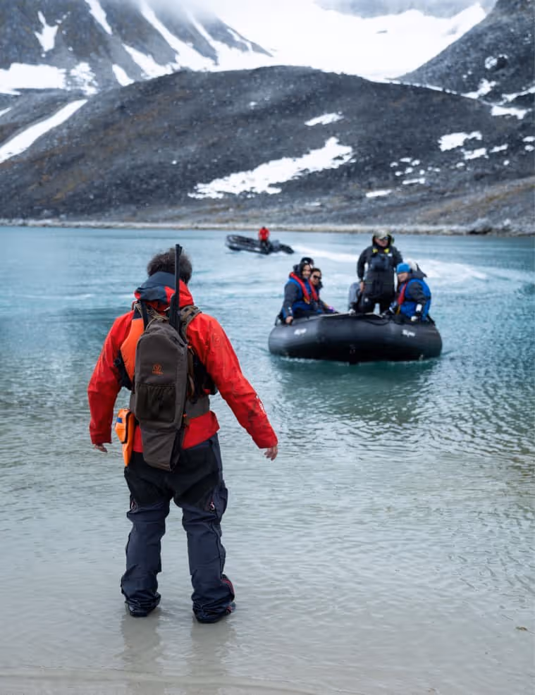 A person in a red jacket standing on the shore, looking towards a small inflatable boat carrying several passengers in a glacial landscape with mountains in the background.