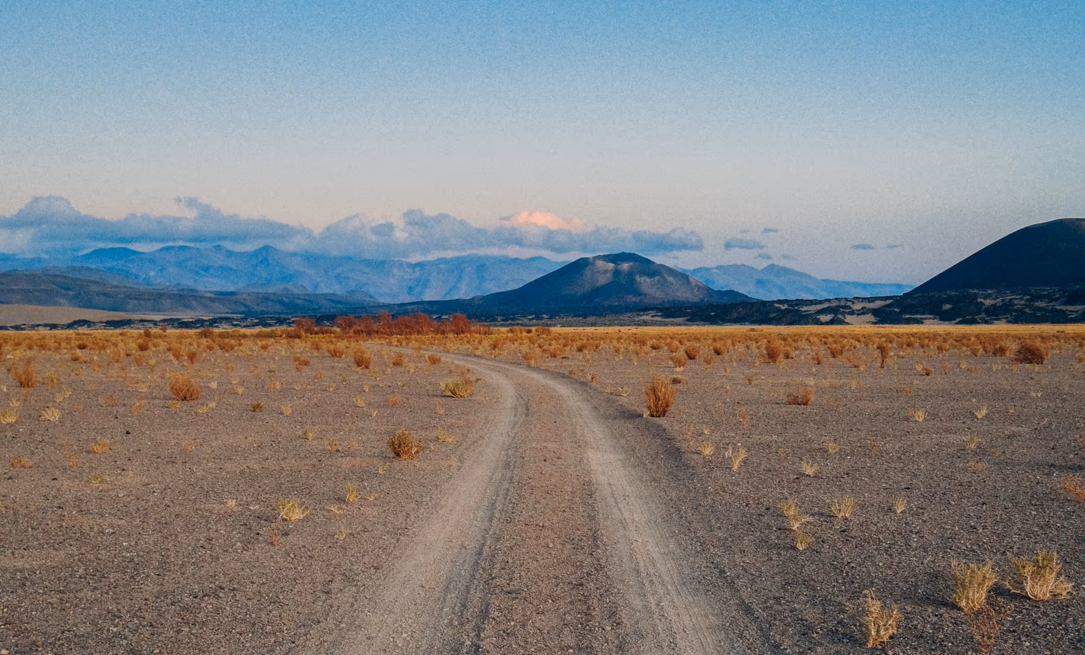 A winding dirt road stretches through a barren landscape with sparse vegetation, flanked by mountains under a clear blue sky. The distant peaks are partially illuminated by the sun.