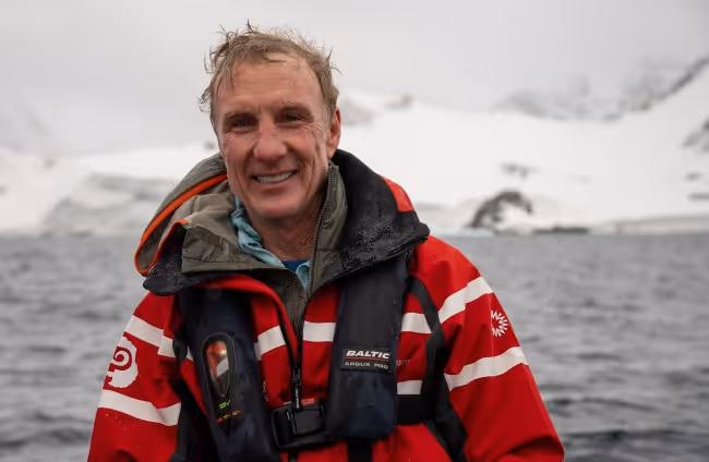 A smiling man wearing a red and black life jacket, standing near icy waters with snow-covered mountains in the background. The scene is overcast, indicating a cold environment.