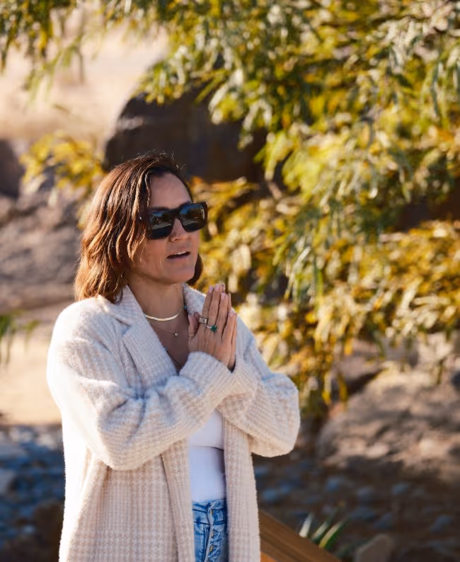 A woman wearing sunglasses and a light sweater stands outdoors, clasping her hands together in a gesture of contemplation or prayer, surrounded by greenery and rocky terrain.