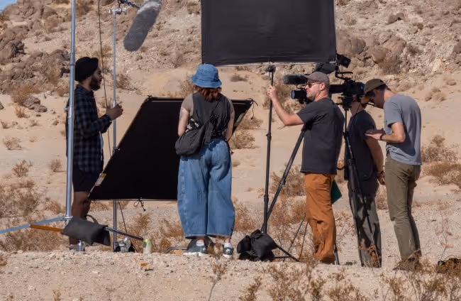 A film crew is working in a desert setting. They are adjusting equipment, including a microphone and reflector, while a woman in denim pants and a hat stands in the center. Two crew members are discussing near a camera.