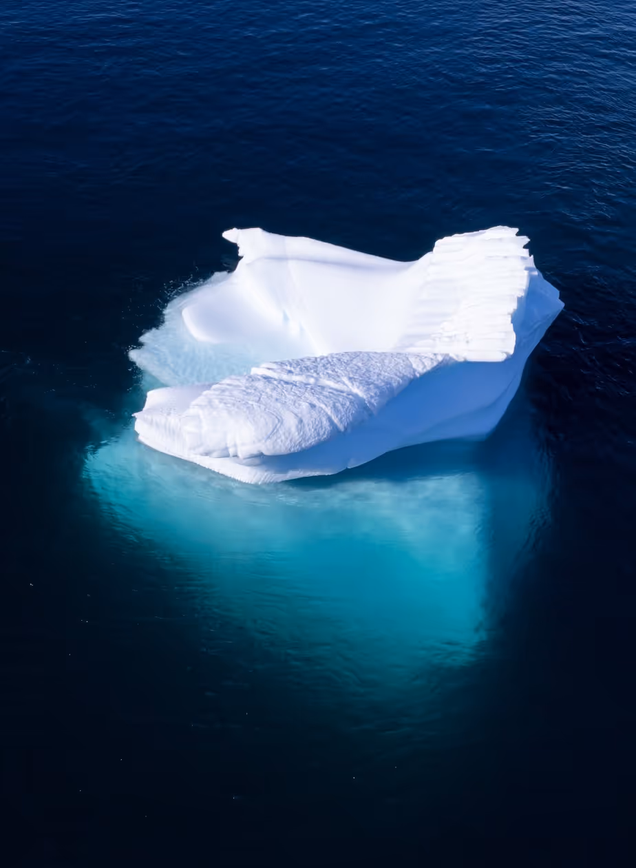 A large, white iceberg floating in deep blue ocean waters, with a smooth, sculptural shape and hints of turquoise beneath the surface.