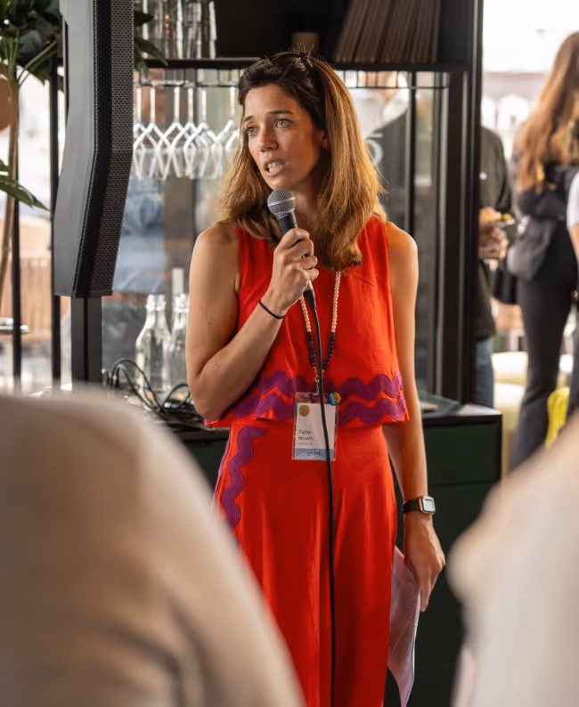 A woman in a red dress speaking into a microphone during an event. She has shoulder-length hair and is standing in front of a bar area with glasses in the background, while audience members are visible behind her.