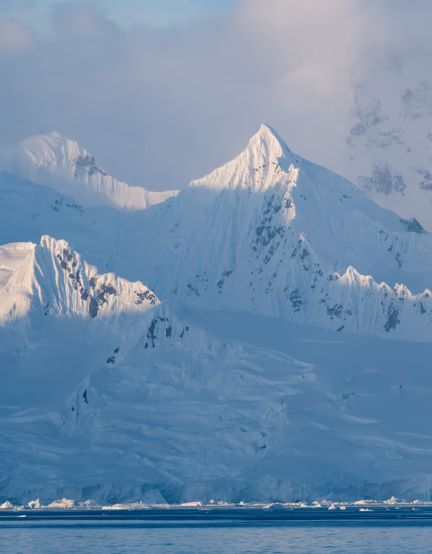 Snow-covered mountains with jagged peaks under a partly cloudy sky, reflected in calm water below. Ice formations are visible at the water's edge.