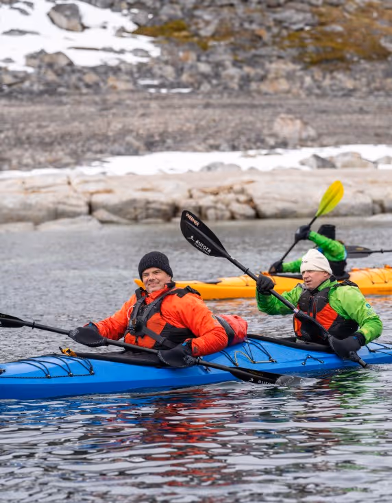 Two people kayaking in a river. One man wearing an orange jacket and black hat is sitting in a blue kayak, while another person in a green jacket paddles nearby. The background features rocky terrain and patches of snow.