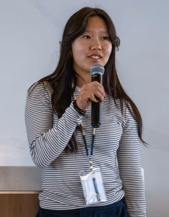 A woman with wavy hair, wearing a striped long-sleeve shirt, speaking into a microphone at an event. She is holding the microphone with one hand and wearing an ID badge on a lanyard.