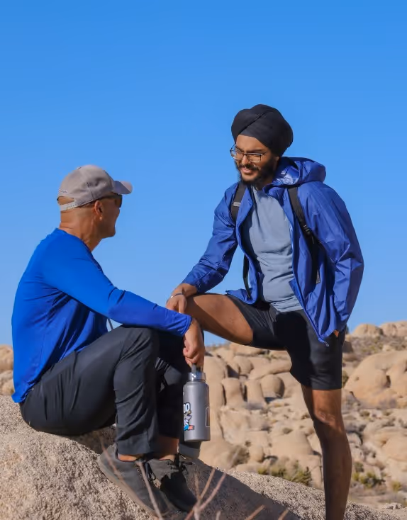 Two men engaging in conversation while resting on a rock formation in a desert landscape. One man wears a blue long-sleeve shirt and cap, and the other sports a blue jacket and a turban.
