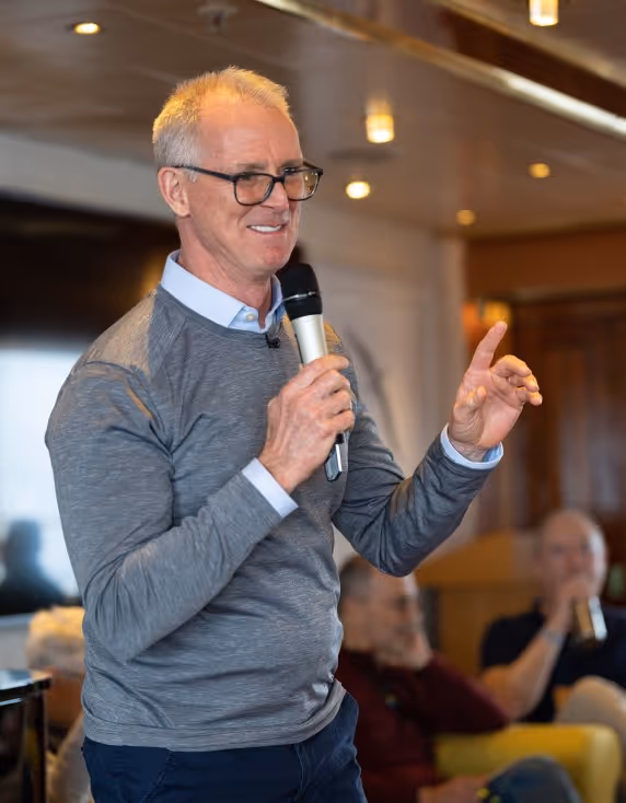 A man with grey hair and glasses speaking into a microphone while gesturing with one hand. He is wearing a grey sweater over a blue shirt in a well-lit indoor setting with blurred audience members in the background.