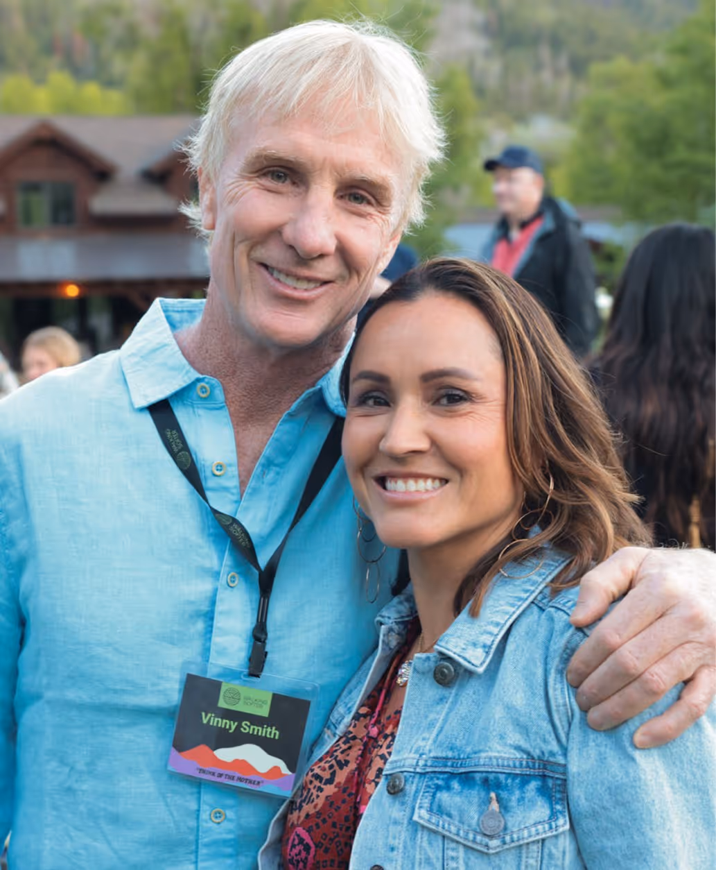 Two smiling individuals posing together outdoors at a social event. One person wears a light blue shirt, while the other has a denim jacket. A name tag is visible on the person in blue.