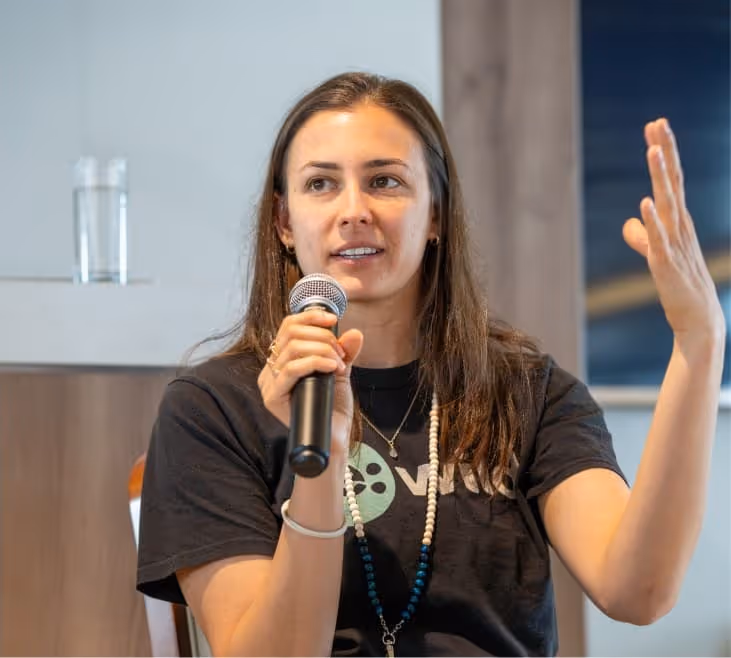 A woman speaking into a microphone while gesturing with her right hand. She is wearing a black t-shirt with a logo, and there is a glass of water on the table behind her.