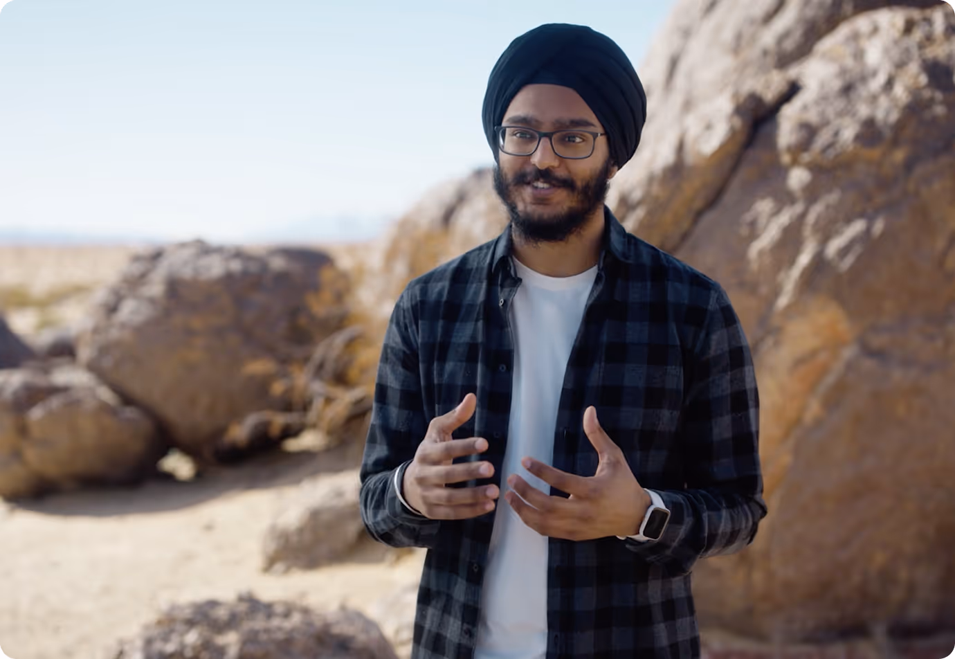 A man wearing a black turban and glasses stands outdoors in front of large boulders, gesturing with his hands while speaking. He is dressed in a checkered shirt and a white t-shirt, with a desert landscape in the background.
