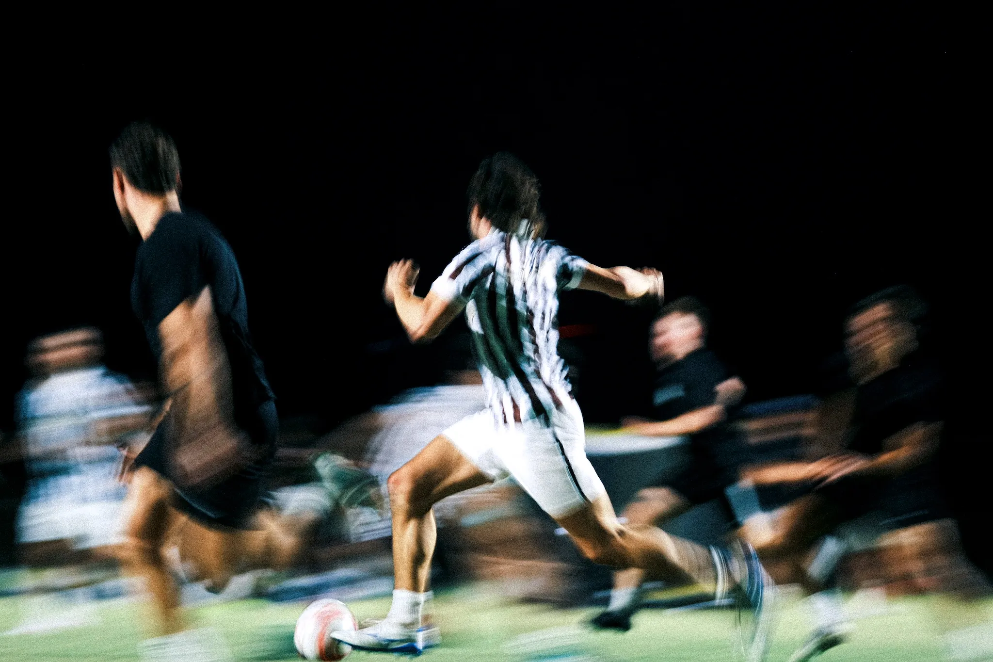 Blurred motion of soccer players running on field at night with one player about to kick the ball.