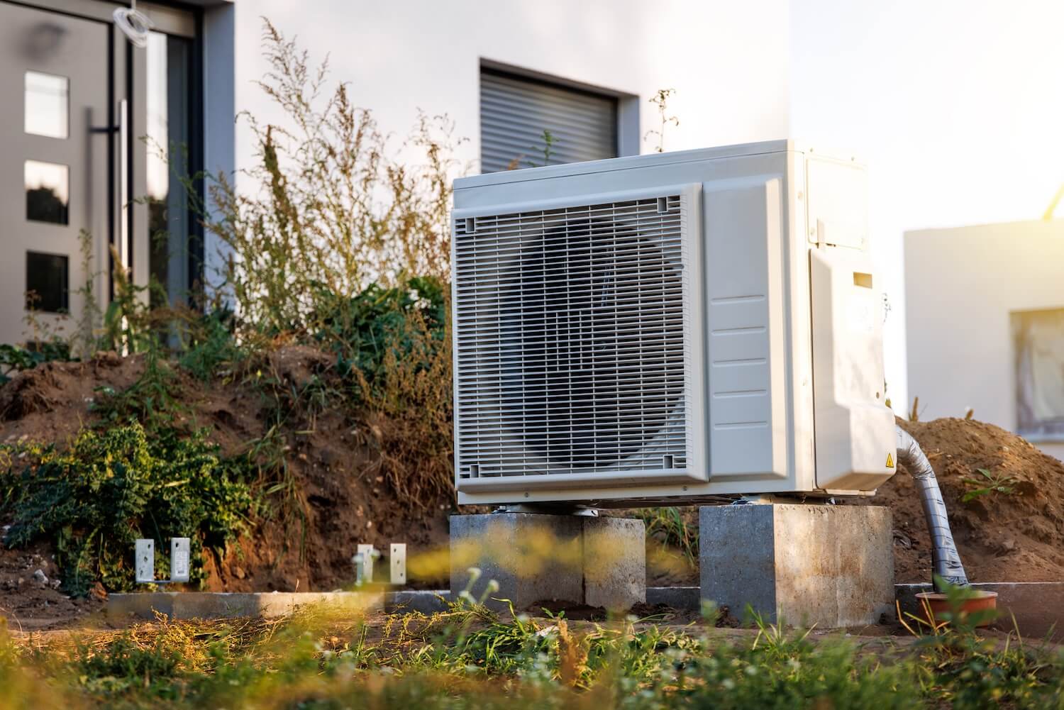 Eine moderne Wärmepumpe im Außenbereich eines Hauses, erhöht auf Betonblöcken platziert – optimal für eine Versicherung der Wärmepumpe.