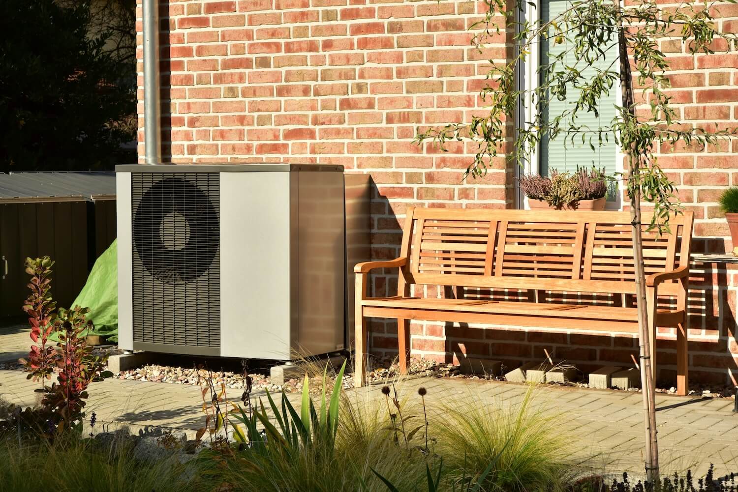 Moderne Wärmepumpe vor einer roten Klinkerwand, eingebettet in einen gepflegten Garten mit Holzbank.