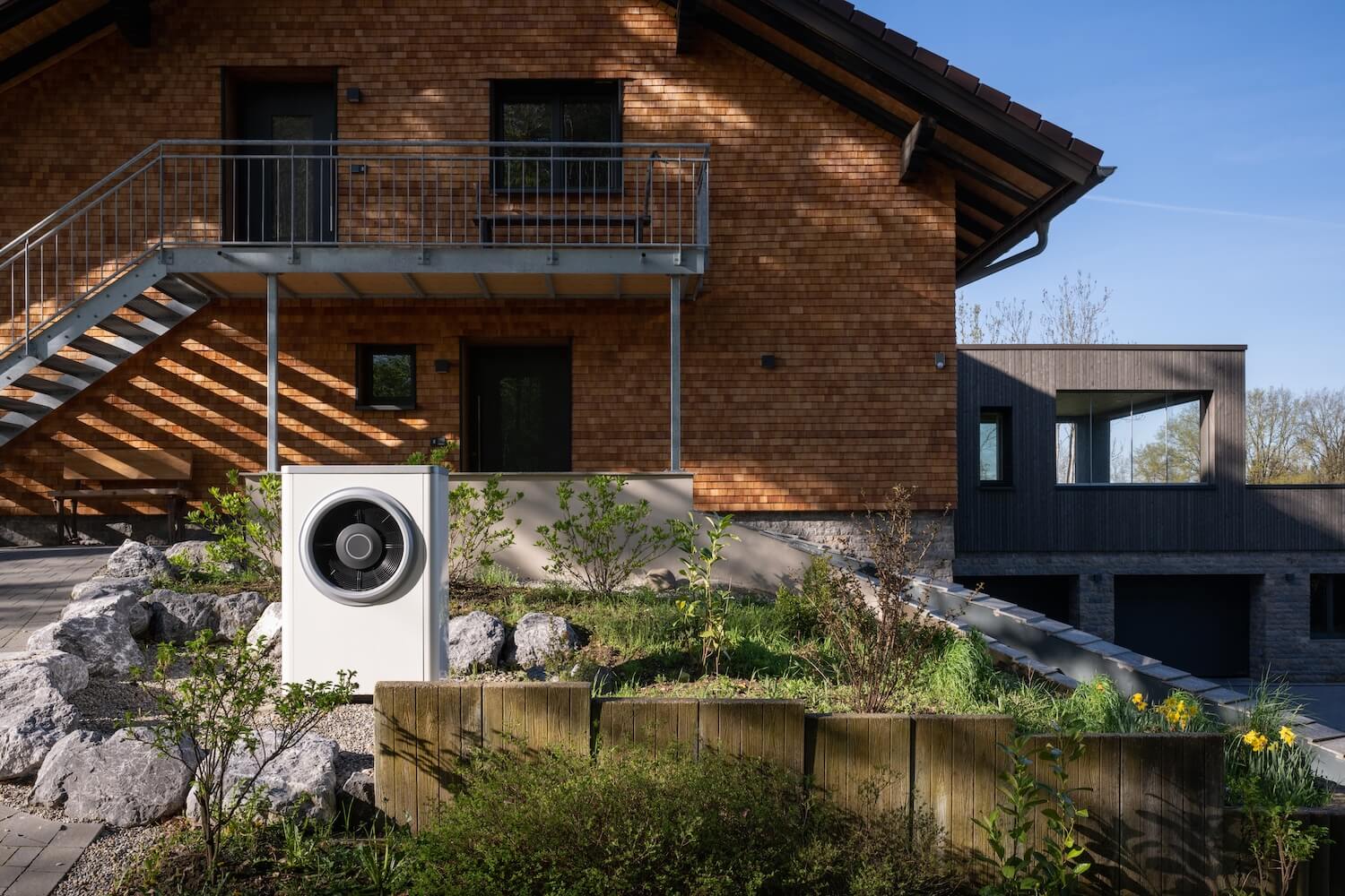 Energieeffiziente Wärmepumpe in einem Garten vor einem Gebäude mit Holzfassade und moderner Architektur.