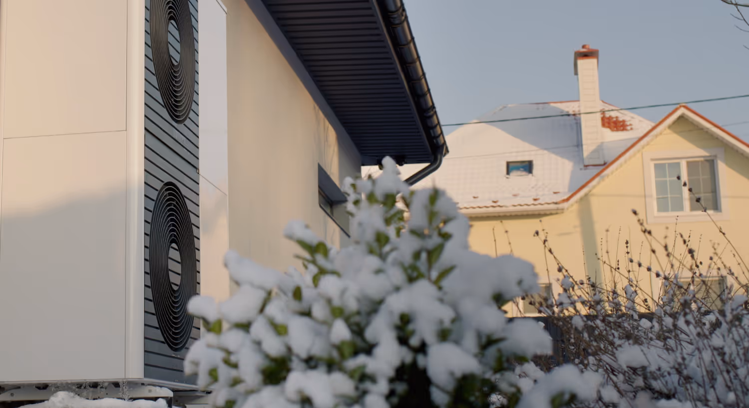 Wärmepumpe vor Hauswand in verschneitem Garten.