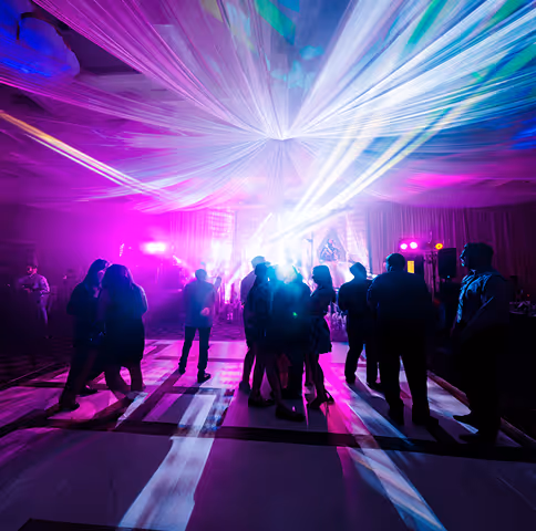 Silhouettes of people dancing on a lit dance floor with vibrant purple and white stage lights and draped ceiling decorations.