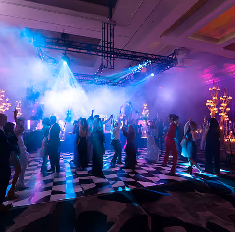 A group of people dancing on a black-and-white checkered dance floor under colorful stage lights and smoke effects in an indoor venue.