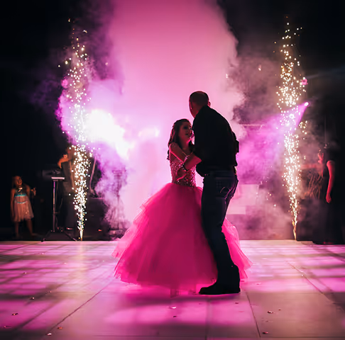 Young woman in a bright pink ball gown dancing with a man on a lit dance floor surrounded by purple smoke and sparklers.