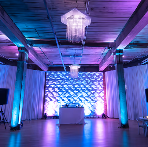 Modern event space with purple and blue lighting, featuring a geometric textured wall, two chandeliers, and a table set for drinks.